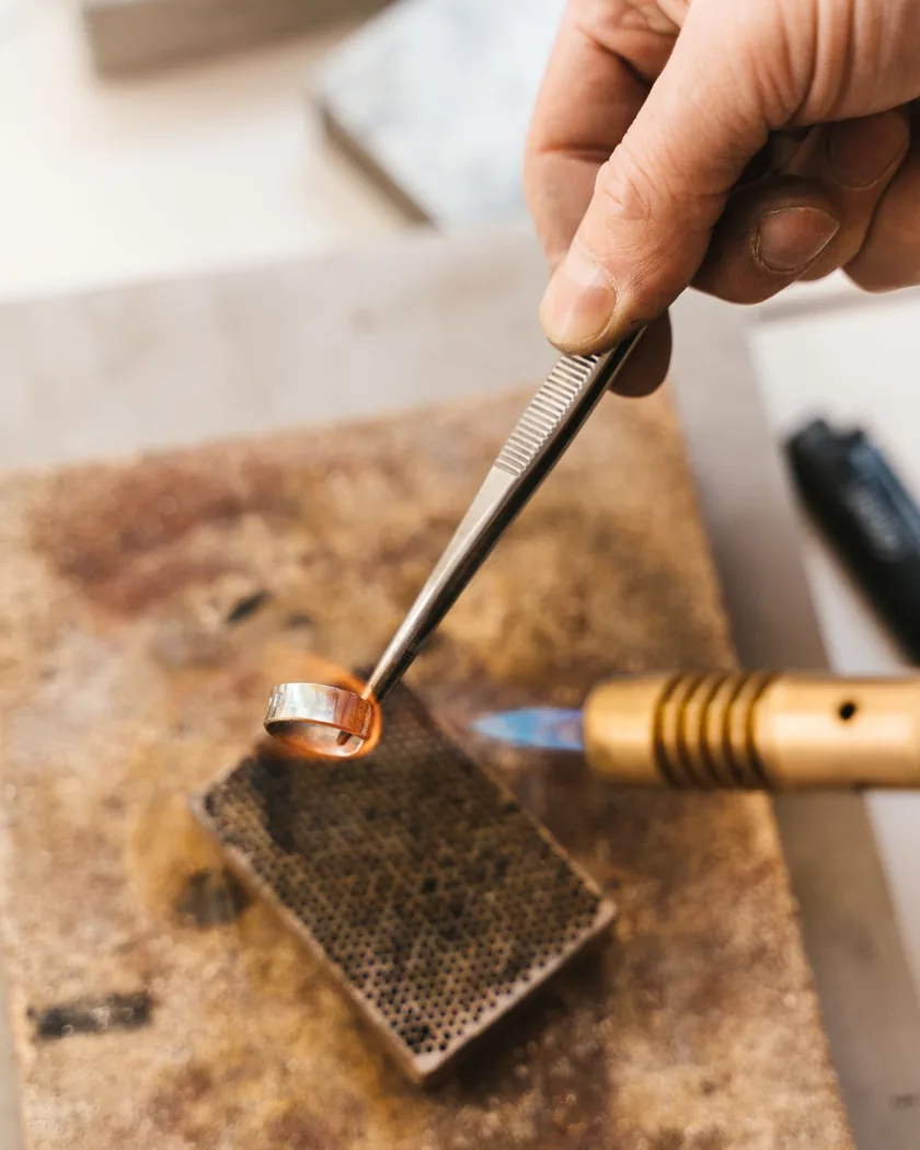 Primer plano de una soldadura de anillo metálico con soplete y pinzas sobre la superficie refractaria del taller.